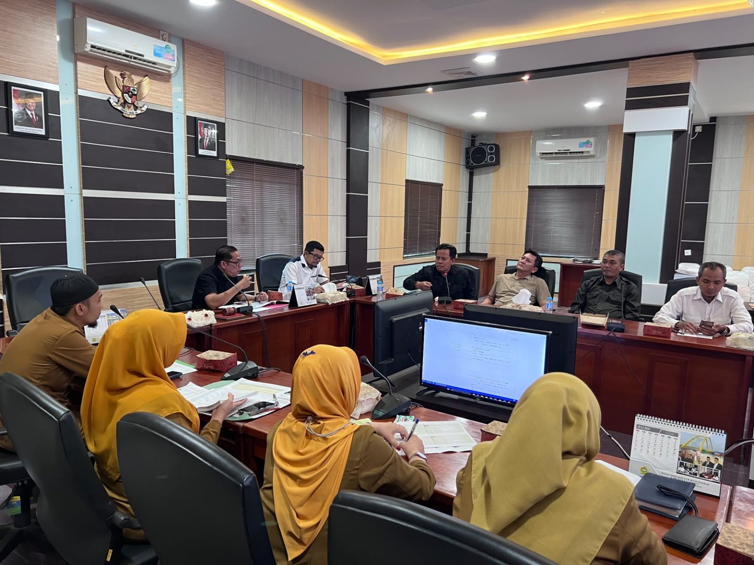 Sudut pandang yang lebih luas memperlihatkan komposisi lengkap peserta rapat. Di satu sisi meja besar, jajaran anggota Komisi I DPRD menyimak presentasi (termasuk legislator dengan kemeja batik biru). Di sisi seberang, delegasi Disdukcapil (termasuk lima petugas berhijab kuning) siap memberikan klarifikasi dan data teknis. Dua layar monitor besar menayangkan substansi rapat – Foto: Sekwan DPRD Karimun