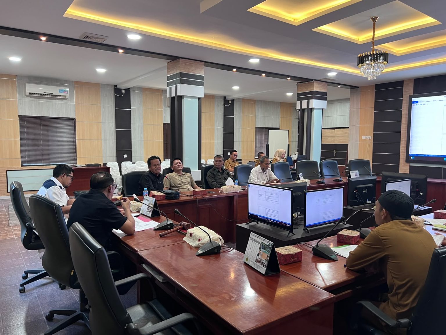 Pengambilan gambar sudut pandang dari ujung meja yang berlawanan, menekankan panjang meja rapat dan partisipasi aktif seluruh peserta. Jajaran tim dewan di sisi kanan dan tim Disdukcapil di sisi kiri, terlibat dalam dialog intensif. Layar monitor menayangkan antarmuka administrasi yang sedang dibahas – Foto: Sekwan DPRD Karimun